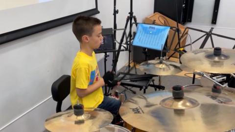 Alfie is pictured on the stage playing the drums. He is wearing a yellow shirt and has a selection of drum symbols in front of him.