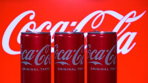 Three Coca Cola cans in a row sitting in front of the large Coca Cola logo