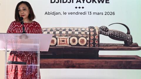 Ivorian Minister of Culture and Francophonie Françoise Remarck talks at a podium in front of a picture of the ornately carved wooden drum