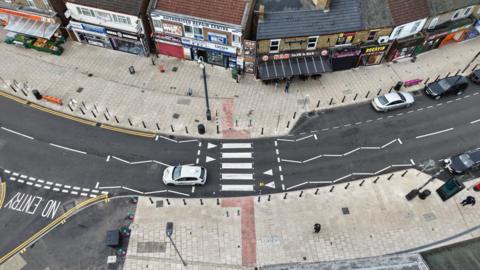 Drone image of the road- with newly painted wite road markings and white pavements seen on either side of it, with cars and people can be seen using the road.