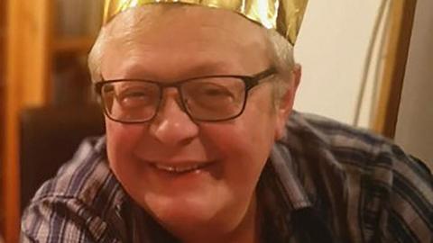Stuart Everett, with grey hair and black-framed glasses, smiles as he poses for a photograph while wearing a gold-coloured paper crown hat.