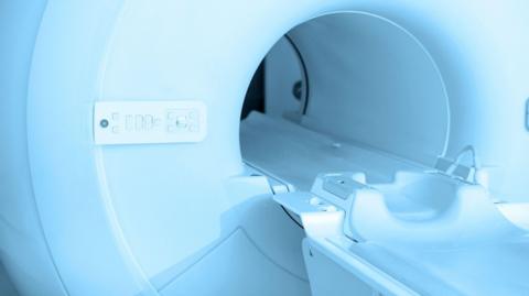 A stock image of a CT canner which shows the entrance to the machine and the bed part where a person lies down. It is all a blueish-white colour and there are buttons and switches on the left of the machine.