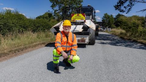 A man in high vis clothing and a work helmet crouches on a road in front of a maintenance vehicle