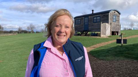 A woman in a pink shirt and navy gilet stands in a field near a wooden cabin.