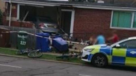A view of a badly damaged house with a silver Mercedes car inside it. The wall of a house has been largely destroyed. There is a police car in the foreground, along with a number of wheelie bins, one lying on its side.