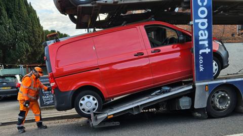 A red van being loaded on to a low loader by a man in a fluorescent orange outfit