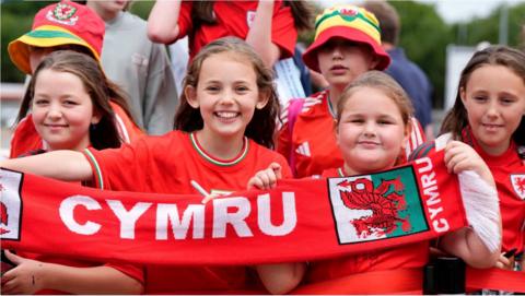 young wales fans wearing red hold a red scarf with the word Cymru