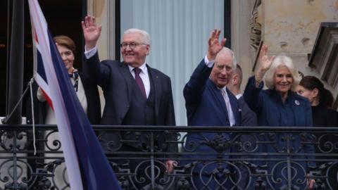 German President Frank-Walter Steinmeier (Centre Left), First Lady Elke Buedenbender (Left), King Charles III and Camilla, Queen Consort, greet crowds from a balcony.