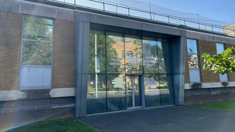 Swansea Magistrates Court, with a bunch of green leaves visible in the right-hand corner. The building is brick with a glass entrance. 