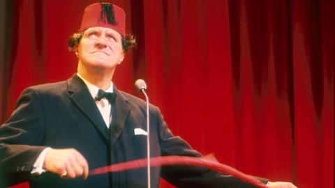 Tommy Cooper, seen here in a fez performing one of his magic tricks. Behind him is a red curtain.