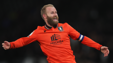 Barry Bannan celebrating after scoring against Derby County, with his arms outstreched. He has light brown hair, a beard, and is wearing a bright orange Sheffield Wednesday away kit.