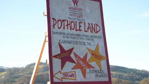 A large handpainted wooden sign with sunlit hills behind. It reads that Wrexham Council proudly presents Pothole Land, with two kilometres of award winning potholes and very little actual road to spoil your fun. 
Guarenteed to be the deepest, longest and widest potholes in Wales.  
The words deepest, longest and widest are displayed in red, orange and yellow star shapes. An arrow with the words, this way, indicates the direction to the left. 