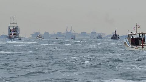 A group of little ships in the water travelling towards the industrial buildings at Dunkirk.