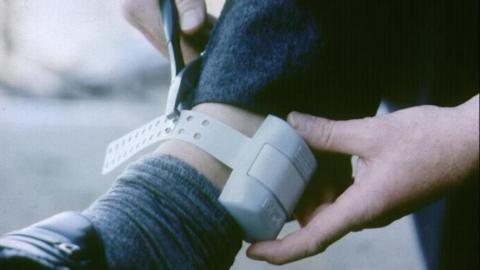 A close-up of a white electronic ankle tag in place with a person's hands holding it.