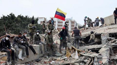 Rescuers search for victims at a collapsed building as one raises a Venezuelan flag following a magnitude 7.2 earthquake that struck Venezuela.