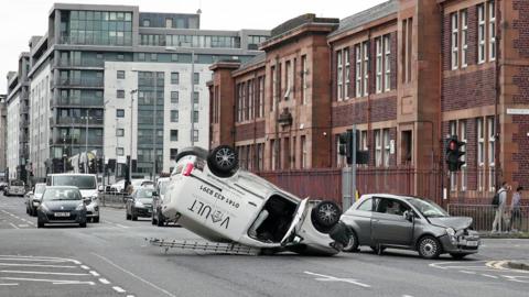 A white van with a ladder on top flipped on it roof with a small grey car with a smashed bonnet beside it.