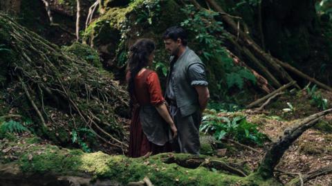 A still of the film Hamnet, showing Jessie Buckley as Agnes and Paul Mescal as William Shakespeare. The pair stand in period dress very close together in the middle of a forest.