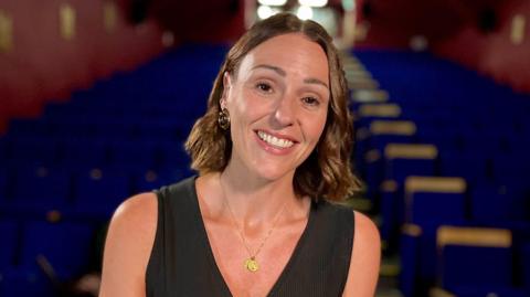 Suranne Jones smiling at the camera, with rows of blue theatre seats behind her. She is wearing a black sleeveless top and a gold coin-like pendant on a thin gold necklace.