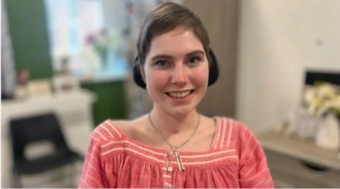 Lily has short brown hair and is wearing headphones. She is wearing a pink top and a gold necklace and is smiling at the camera