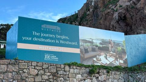 A picture of a sign with a brick wall below it. The sign has an image of a property on it, next to writing. It is blue in colour.