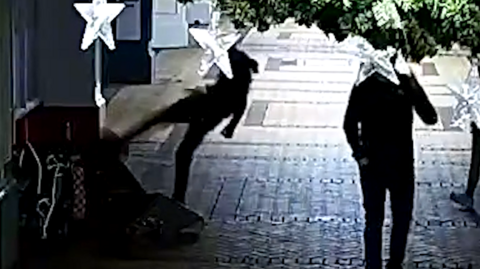 CCTV of three people standing in a street. One man is shown kicking someone on the floor. There are items to the left of the image but it is not clear what they are. The faces of the people are not revealed. There are buildings to the left. 