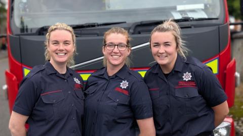 L-R Ciara McGurk, Rachael McKeown, Michaela McGuigan. All three are wearing their navy NIFRS uniforms and are standing near a fire engine. 