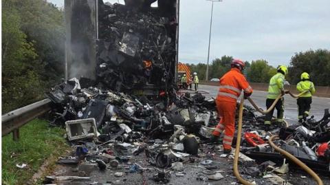 Firefighters battle the flames of a lorry fire on the M5 in Exeter. Two crew members are inspecting breathing apparatus on the road while others work on the fire.