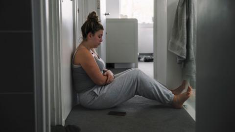 A young woman sits in the hallway of her home looking sad and lonely