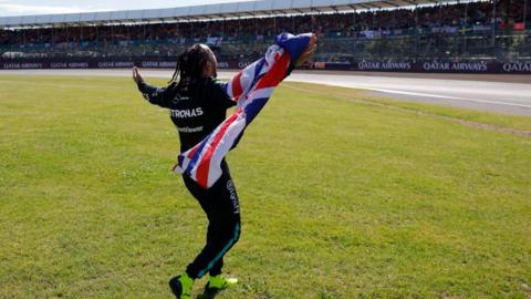 Lewis Hamilton in black racing uniform waving a union flag at a stand at Silverstone