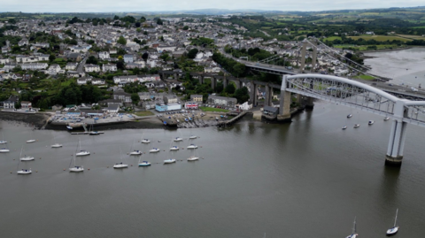 Aerial view of two bridges spanning a river with boats moored in the river and houses on the bank of the river with fields beyond.