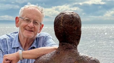 Ron Davies with grey hair wearing a blue and white shirt and glasses leans on one of the Iron Men sculptures on Crosby beach with the incoming tide in the background on a clear day. He is smiling. 