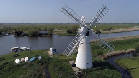 Thurne Windmill complete with sails on the banks of the River Thurne, Norfolk Broads. Grassy banks, a few boats in the water, some upturned canoes on the grass banks. The land is very flat and the waterway is still. 