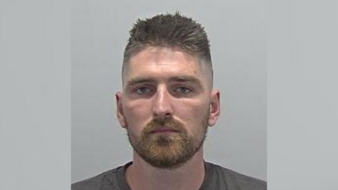 A mugshot image of Alex Stokes. He is wearing a dark grey t-shirt and standing against a grey wall