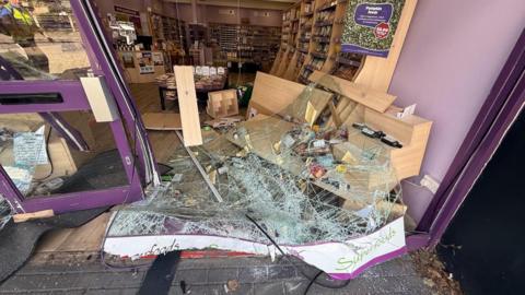 The picture shows the front of a store that has been destroyed. There is broken glass on the floor, the doors are bent inwards, shelves have fallen along with the goods in them. The back of the store is completely untouched, as products can be seen lined up neatly on shelves.