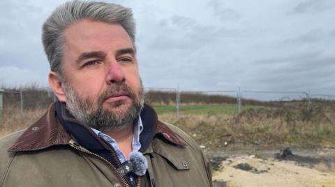 A man with slicked back grey hair and a matching beard stands in front of a fenced-off piece of green land under a grey sky. He is wearing a brown jacket over a blue shirt.