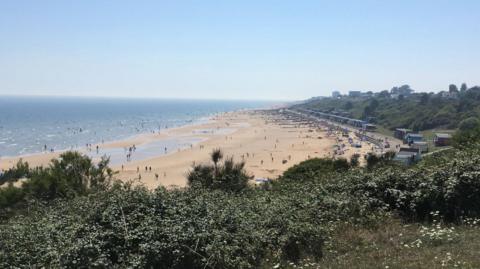 A wide expanse of sandy beach with dozens pictured from a cliff top. The edge of the cliff is covered in bushes. The camera is pointed along the beach, with blue sea on the left, about 100m of beach, and a prom, groynes, beach huts and bush-covered earthen cliff on the right. Tall building are visible on the horizon.