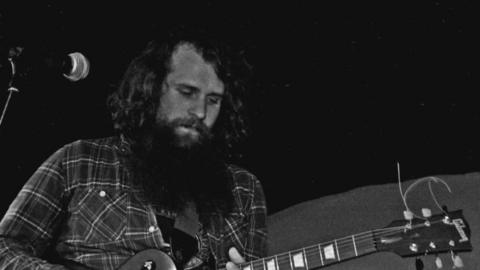 A black and white photo of a man with a long beard and long curly hair playing a guitar while wearing a checked shirt. 