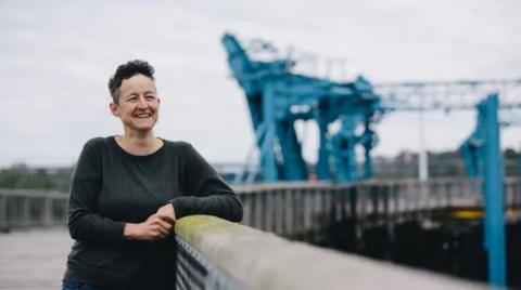 Kari Vickers leaning on a timber railing at Dunston Staiths. She has short dark hair and is smiling as she looks into the distance. The staiths is a timber structure. A metal clamp can be seen in the background, stabilising part of the wood.