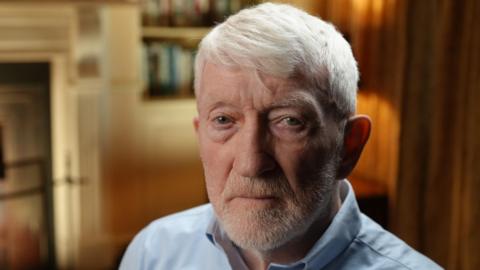 Brendan Hughes looks directly into the camera. He has grey hair and a short beard with a mix of green and brown in his eyes. Brendan is wearing a blue shirt with the top button open. Behind him is an out of focus room with long curtains, a fireplace and a bookshelf with warm lighting which gives off an orange glow. 