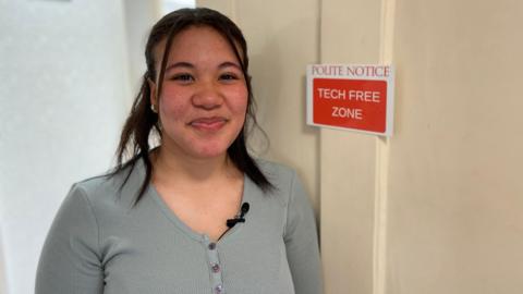 A teenage girl smiles in front of her bedroom door, which has the sign 'Polite notice: Tech free zone'
