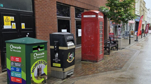 A red phone box on a street in Worcester. There is a black metal bench on the right of it and recycling and litter bins on the left. There are shops, signs and a small tree in the background