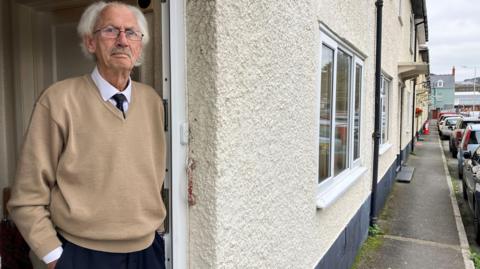 Picture of a 79 year old man with silver hair and glasses, leaning on his front door frame, wearing a shirt, tie and jumper. 