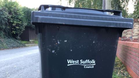 A general view of a West Suffolk black waste bin on a pavement. A road passes next to it.