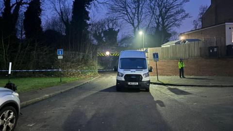 A railway bridge is cordoned off with blue and white police tape. A police officer is standing by the cordon in uniform. There is a white van parked in front of the cordon.