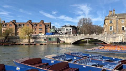 Punts lined up in the river with a bridge and buildings behind and bright blue sky 