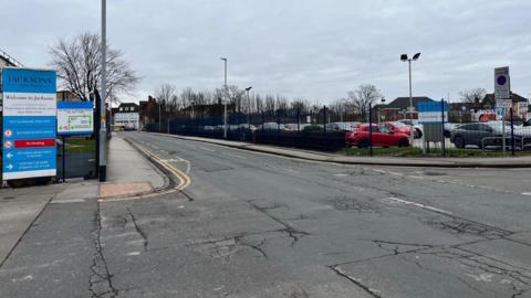 An image of Derringham Street in Hull taken near the entrance to Jacksons Bakery. The road is cracked and has several potholes. The junction with Spring Bank is visible in the distance. A car park is on the right of the image.