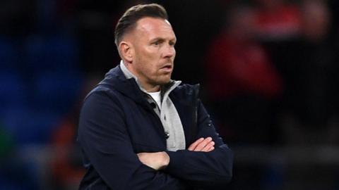 Craig Bellamy, in an open dark jacket with a grey top underneath, looks on with a focused stare during Wales' match against Northern Ireland