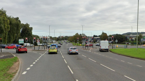 A busy junction controlled by traffic lights. A number of cars are waiting at the lights. A retail park is visible to the right of the shot.