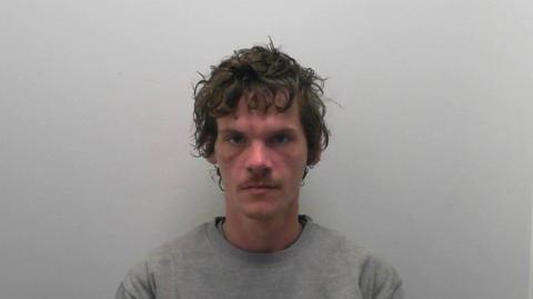 Daniel Bell in a police mugshot. He has medium length brown hair and a small brown moustache and is wearing a grey sweatshirt