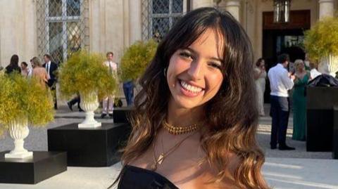 Grace O'Malley-Kumar smiling for the camera with long brown hair while holding a glass of wine at an outdoors event.